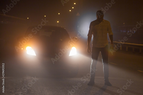 Incident on the road. Unknown person stands in the middle of the roadway at night in the fog by the headlights. Concept of the attack on the road