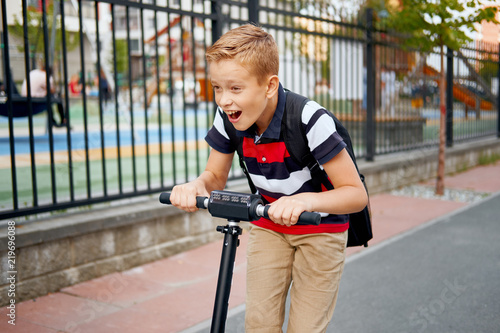 School boy in riding with his electric scooter in the city with backpack on sunny day. Child in colorful clothes biking on way to school