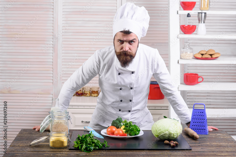 Bearded serious chef stands in kitchen near table. Cook in white uniform stands on restaurant kitchen. Master chef. Cooking, culinary, cuisine. Vegetarian, health, meal, diet. Food preparation concept Bearded serious chef stands in kitchen near table. Cook in white uniform stands on restaurant kitchen. Master chef. Cooking, culinary, cuisine. Vegetarian, health, meal, diet. Food preparation concept