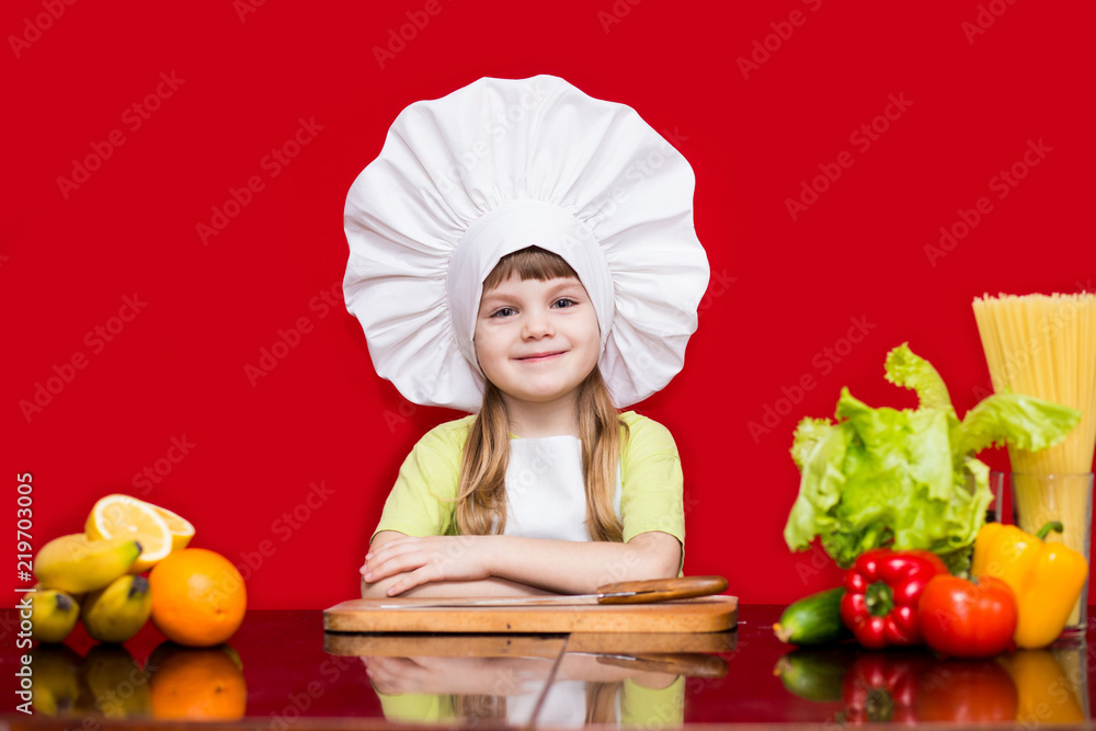 Happy little girl in chef uniform cuts fruit in kitchen. Kid chef. Cooking Process Concept Happy little girl in chef uniform cuts fruit in kitchen. Kid chef. Cooking Process Concept