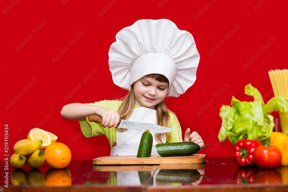 Happy little girl in chef uniform cuts vegetables in kitchen. Kid chef. Vegetarian. Cooking Process Concept Happy little girl in chef uniform cuts vegetables in kitchen. Kid chef. Vegetarian. Cooking Process Concept