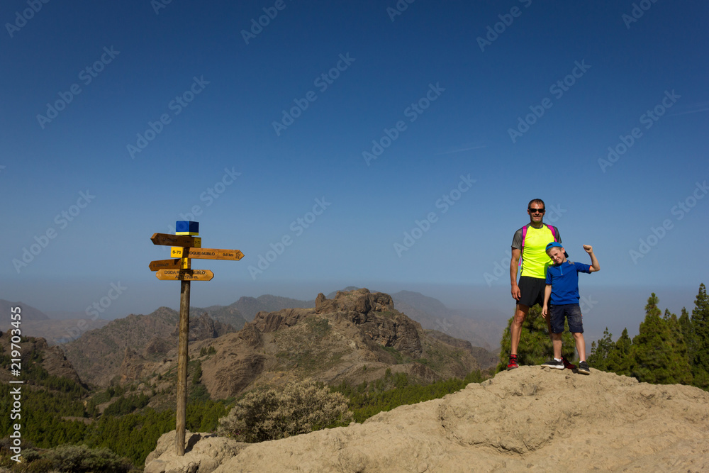 Dad and little boy standing on top of mountain by hiking signs on sunny day in Roque Nublo, Gran Canaria. Family outdoors activity, healthy lifestyle, climb, explore, adventure concepts Dad and little boy standing on top of mountain by hiking signs on sunny day in Roque Nublo, Gran Canaria. Family outdoors activity, healthy lifestyle, climb, explore, adventure concepts