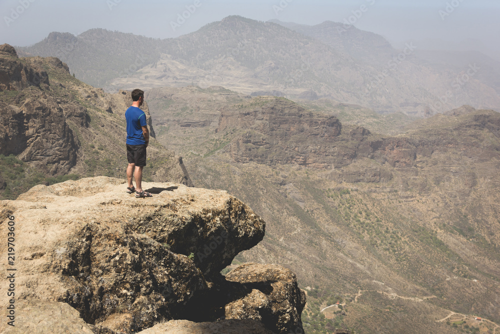 Fototapeta premium Tourist on mountain edge facing great landscape views. Young man on blue t shirt standing on top of rock on sunny day in Roque Nublo. Adventure, explore, freedom, visionary, solitude concepts