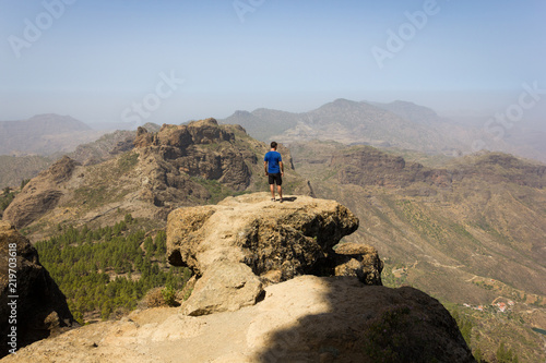 Young man on blue shirt standing on rock top on sunny day in Roque Nublo, Gran Canaria. Hiker on mountain edge facing panoramic views. Outdoor activity, adventure, explore, freedom concepts