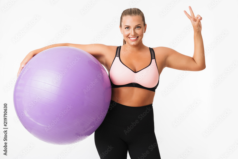 Pretty smiling plump girl in sporty top holding fitness ball in hand happily showing two fingers gesture while looking in camera over white background. Plus size model Pretty smiling plump girl in sporty top holding fitness ball in hand happily showing two fingers gesture while looking in camera over white background. Plus size model