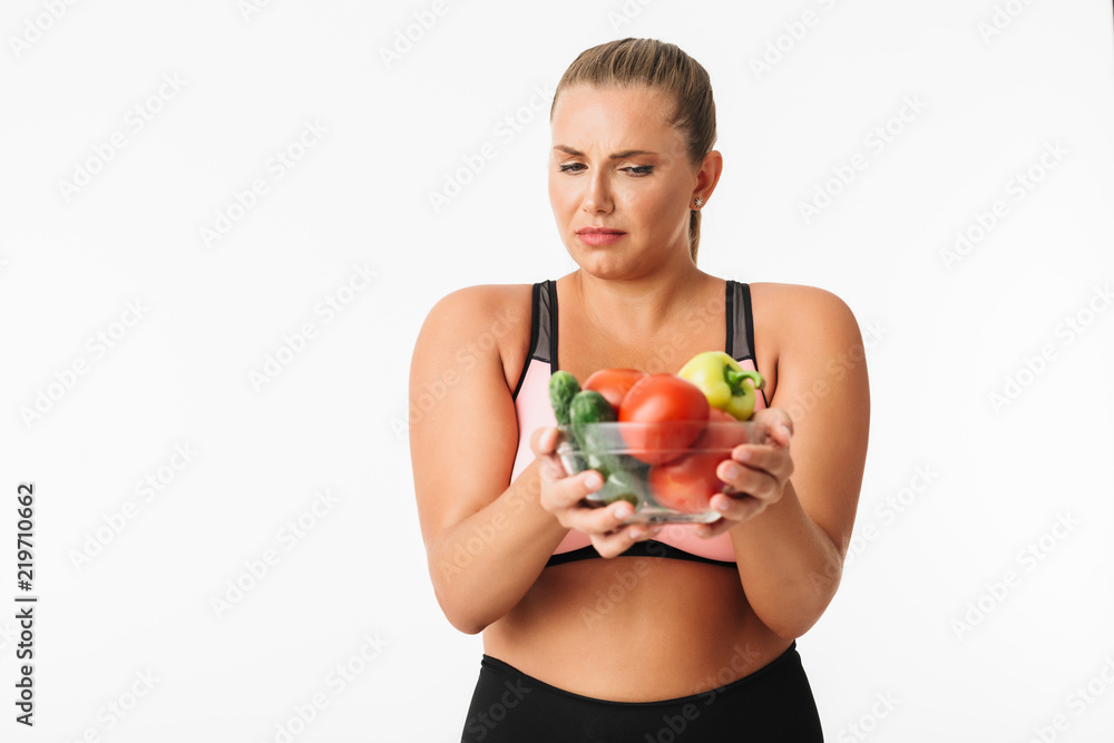 Young disappointed woman with excess weight in sporty top sadly looking on bowl with vegetables in hands over white background isolated. Plus size model Young disappointed woman with excess weight in sporty top sadly looking on bowl with vegetables in hands over white background isolated. Plus size model
