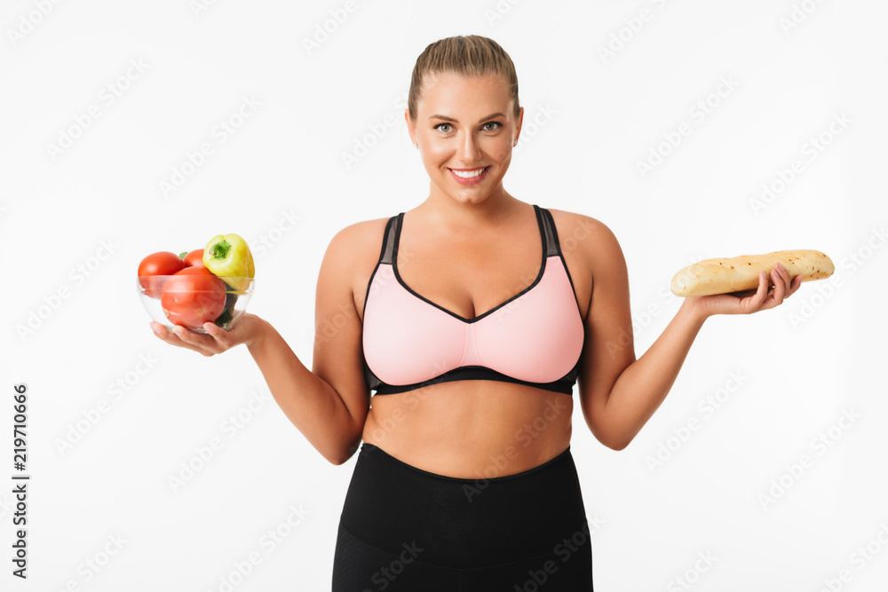 Smiling girl with excess weight in sporty top holding bowl with vegetables and bread in hands while joyfully looking in camera over white background. Plus size model Smiling girl with excess weight in sporty top holding bowl with vegetables and bread in hands while joyfully looking in camera over white background. Plus size model