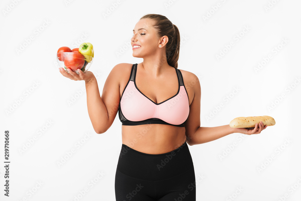 Cheerful girl with excess weight in sporty top happily holding bowl with vegetables and bread in hands over white background isolated. Plus size model Cheerful girl with excess weight in sporty top happily holding bowl with vegetables and bread in hands over white background isolated. Plus size model