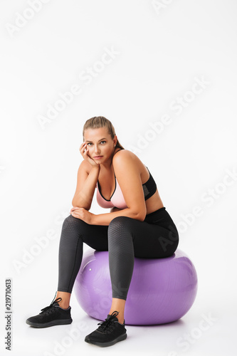 Beautiful woman with excess weight in sporty top and leggings sitting on fitness ball while thoughtfully looking in camera over white background. Plus size model