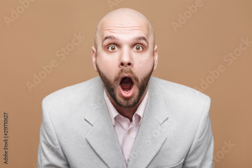 Pointing finger and amazed portrait of handsome middle aged businessman in classic light gray suit standing, looking at camera, wondering and showing brown background. indoor studio shot, isolated.