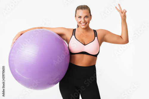 Pretty smiling plump girl in sporty top holding fitness ball in hand happily showing two fingers gesture while looking in camera over white background. Plus size model