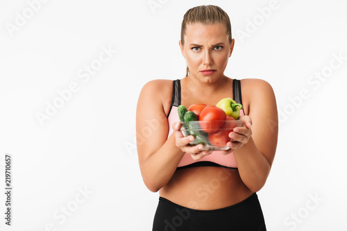 Young woman with excess weight in sporty top sadly looking in camera while holding bowl with vegetables in hands over white background. Plus size model © Anton