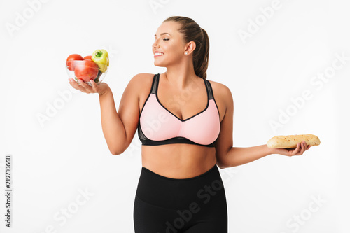 Cheerful girl with excess weight in sporty top happily holding bowl with vegetables and bread in hands over white background isolated. Plus size model © Anton