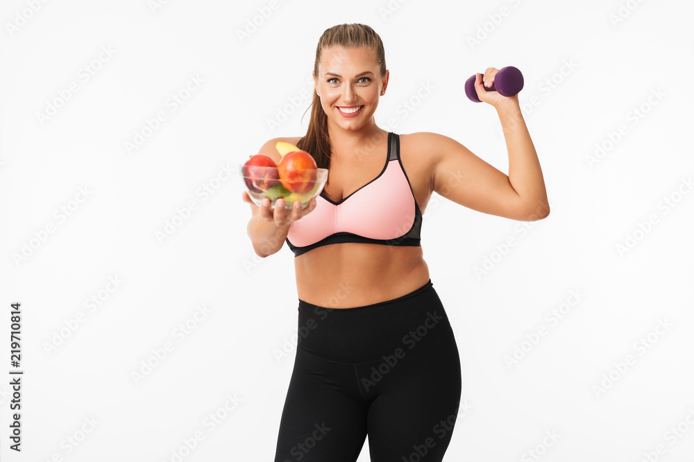 Smiling girl with excess weight in sporty top holding dumbbell in hand while happily showing bowl with fruits on camera over white background. Plus size model Smiling girl with excess weight in sporty top holding dumbbell in hand while happily showing bowl with fruits on camera over white background. Plus size model