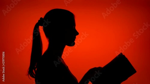 Silhouette of young woman reading physical print book on red background. Female's face in profile studying. Black contur shadow of teenager's half-face. Concept of old traditional classical paper
