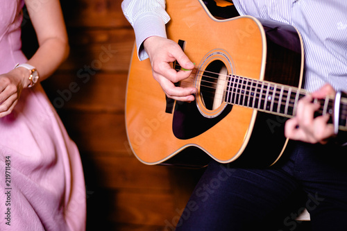 Guitar with a man's male hands playing the guitar near a women singer pink dress on wooden wall background, acoustic guitar with nature light. Concept of guys boys jazz band performing on events . 