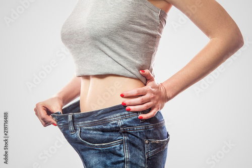 Closeup of slim waist of young woman standing in big jeans and gray top showing successful weight loss, indoor studio shot, isolated on light gray background, diet concept.