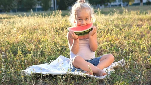 A cute little blonde is sitting in the park on the grass and gaily eating a watermelon.