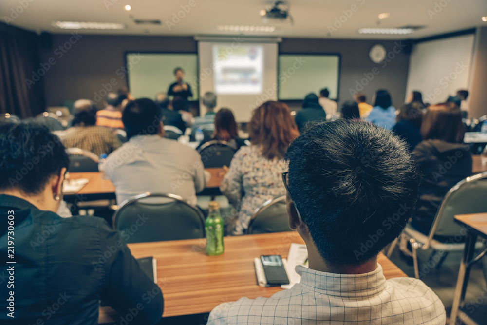 Back side of audience listening the Asian Speaker with casual suit on the stage in front of the room in the conference hall, business and education concept