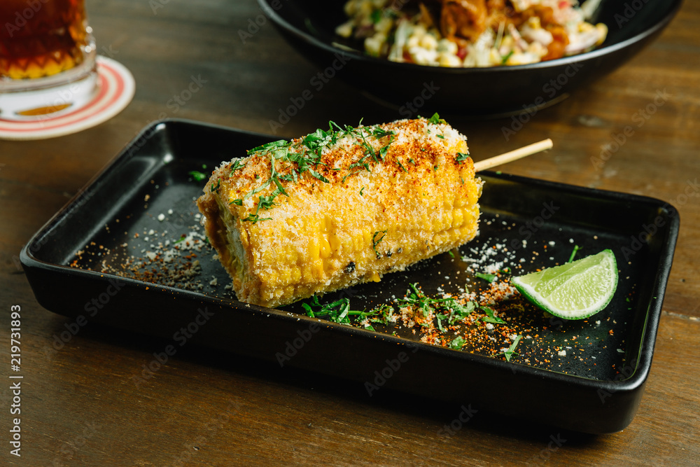 Mexican style grilled corn topping with cheese and chili powder served with sliced lime in black plate. Mexican style grilled corn topping with cheese and chili powder served with sliced lime in black plate.