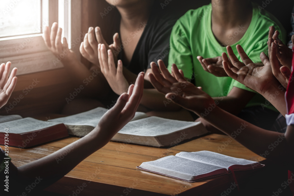Obraz premium christian children group praying together around wooden table with bible in home room, prayer meeting concept