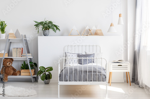 Gray bedding on a single bed with metal frame and a scandinavian style nightstand in a beautiful, bright kid bedroom interior with natural light