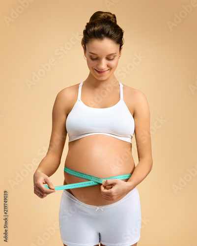 Attractive pregnant woman with measuring tape measures her belly on beige background. Pregnancy, maternity, preparation and expectation concept © Romario Ien