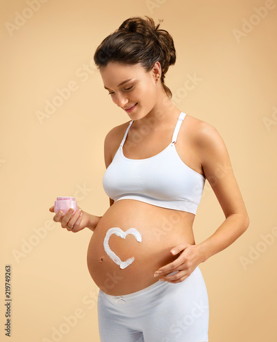 Smiling pregnant woman applying moisturizing cream on her belly on beige background. Pregnancy, maternity, preparation and expectation concept