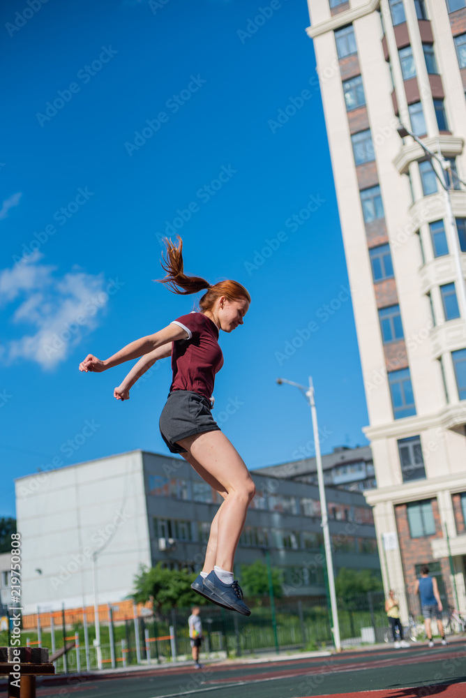 the red-haired student in shorts gives the standards for jumping at the physical education class on an open sports ground. girl doing crossfit exercises outdoor. red-haired young woman is jumping