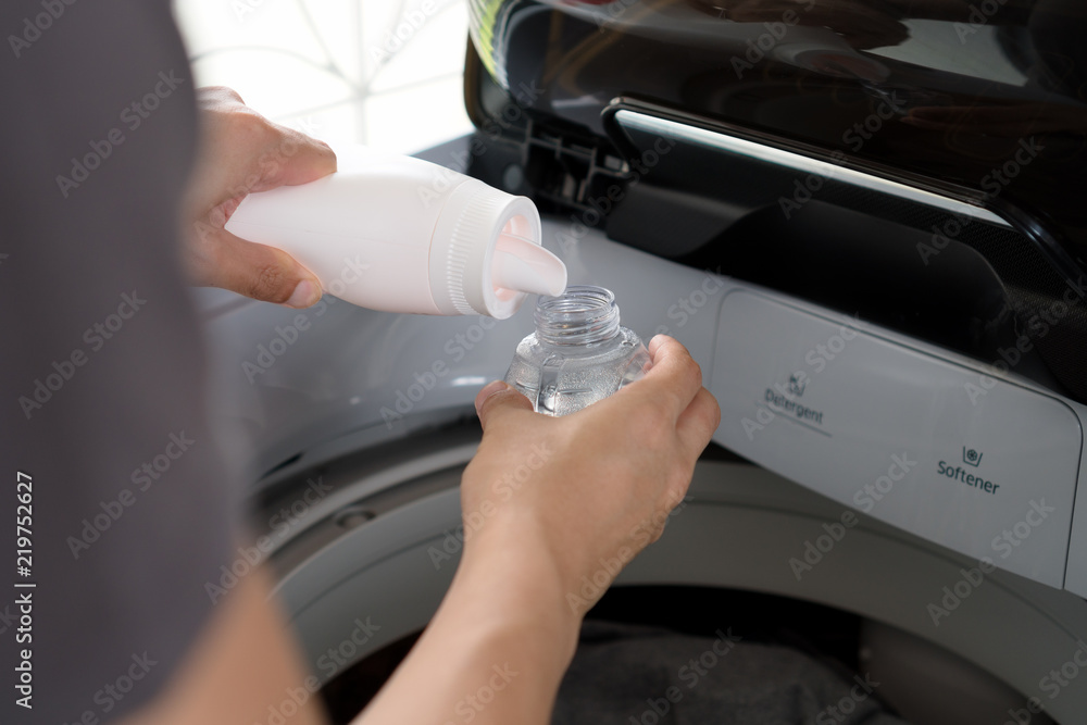 Man hand pouring liquid laundry detergent and softener in bottle cap with washing machine.
