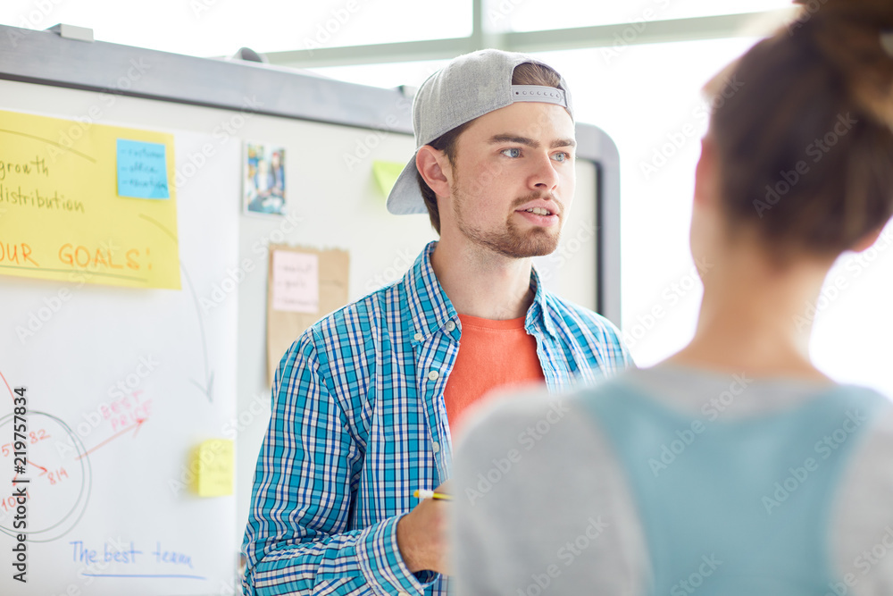 Confident enterprising student boy with beard discussing project with groupmate and presenting his idea in modern classroom with whiteboard Confident enterprising student boy with beard discussing project with groupmate and presenting his idea in modern classroom with whiteboard