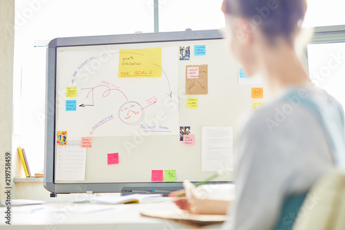 Defocused woman sitting at table and learning information on whiteboard while sitting in meeting room with marketing plan on whiteboard