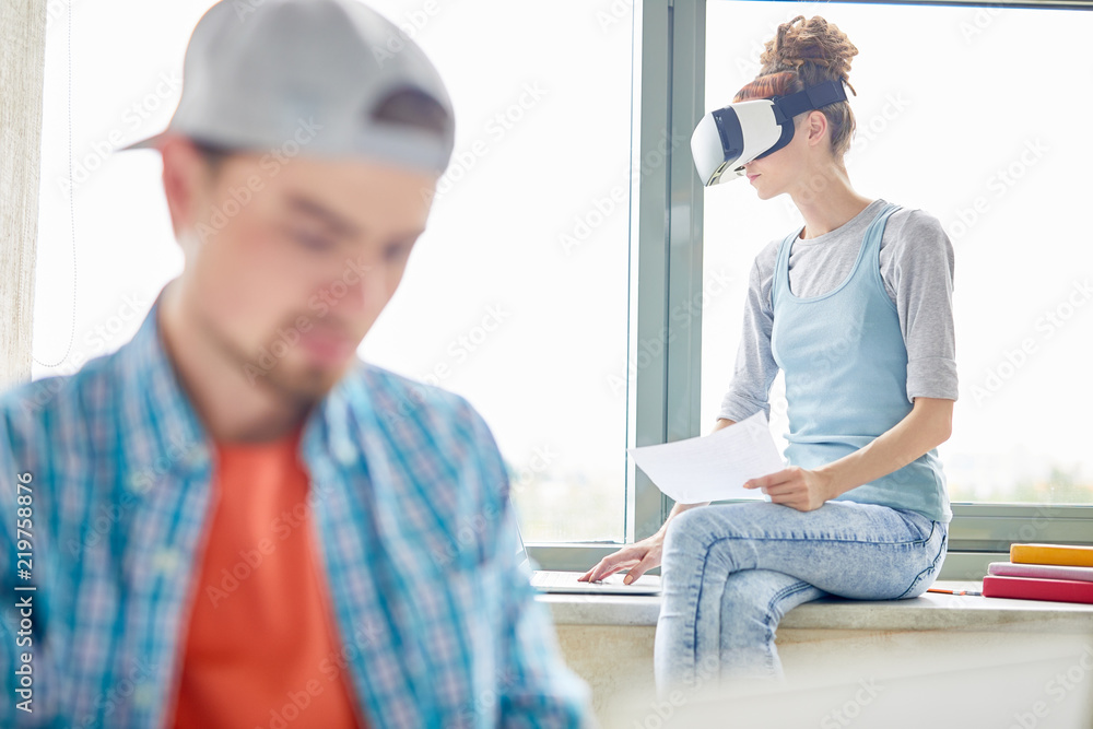 Fototapeta premium Pensive young female student in VR headset sitting on window-sill and using laptop while examining data, she working on project