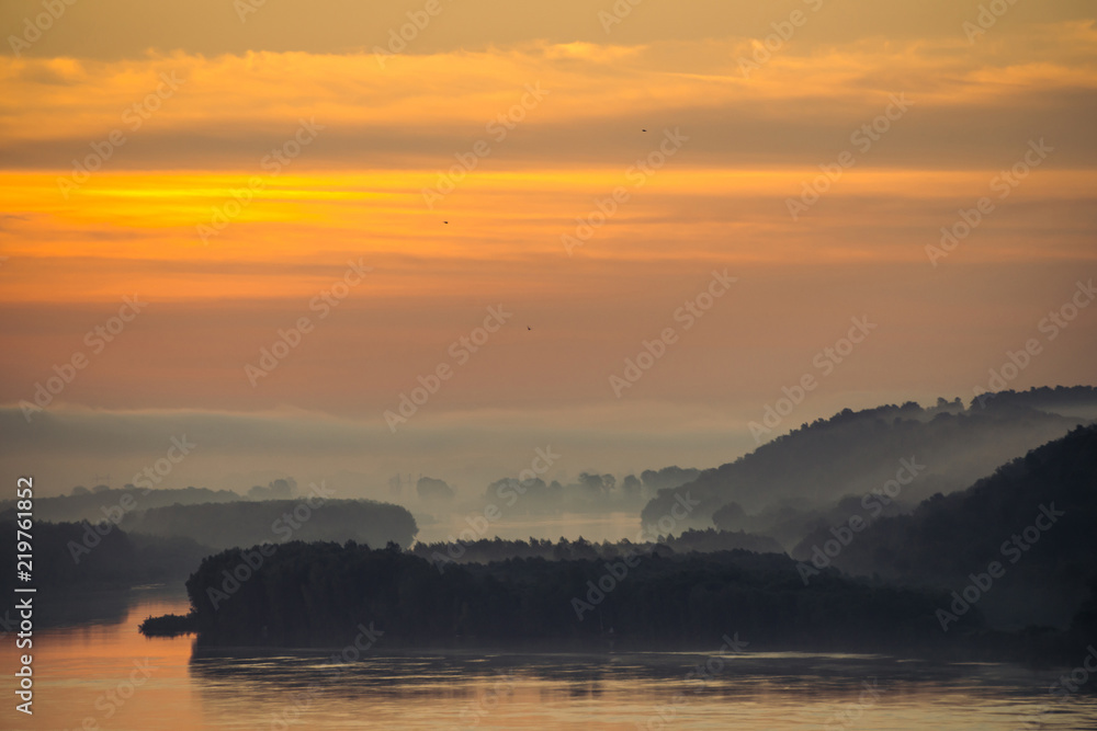 Morning haze above valley of river. Gold glow from dawn in sky and reflex on water. Birds flying in sky at sunrise. Fog on riverbank with forest. Colorful atmospheric landscape of majestic nature.