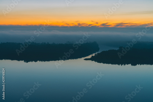 Fototapeta Naklejka Na Ścianę i Meble -  Early blue sky reflected in river water. Riverbank with forest under predawn sky. Yellow stripe in picturesque sky. Fog hid trees on island. Mystical morning atmospheric landscape of majestic nature.