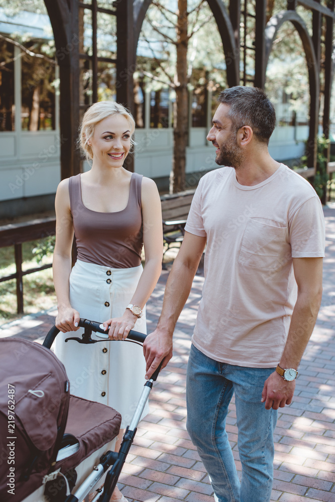 smiling parents walking with baby carriage in park and looking at each other smiling parents walking with baby carriage in park and looking at each other
