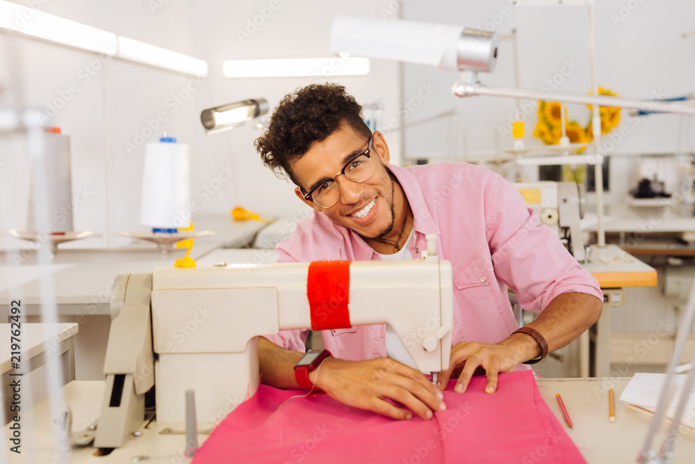 Nice workplace. Cheerful enthusiastic young man smiling and feeling good while sewing pink textile on the sewing machine Nice workplace. Cheerful enthusiastic young man smiling and feeling good while sewing pink textile on the sewing machine