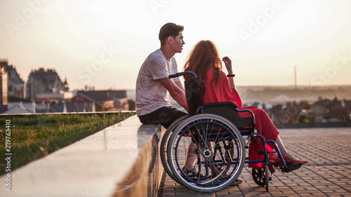 Konstfotografi Young couple admiring the sunset, the girl in a wheelchair, the guy sitting next