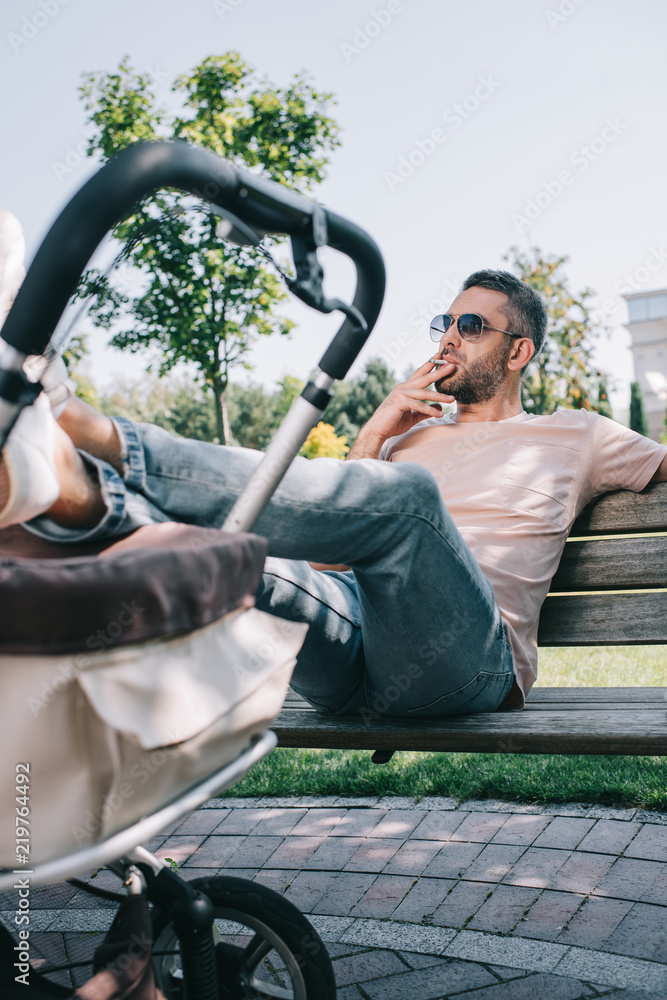 Obraz premium father smoking cigarette on bench with legs on baby carriage in park