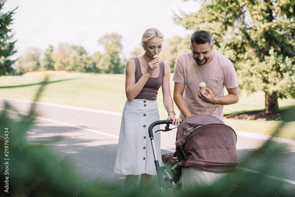 parents eating ice cream and looking at baby carriage in park parents eating ice cream and looking at baby carriage in park