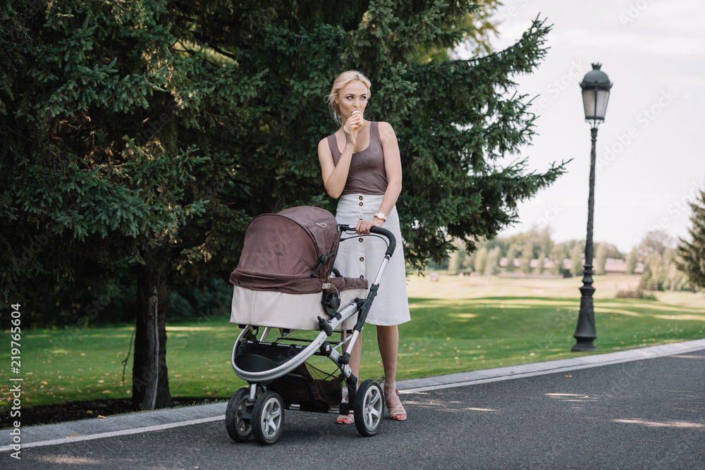 mother walking with baby pram and eating ice cream in park