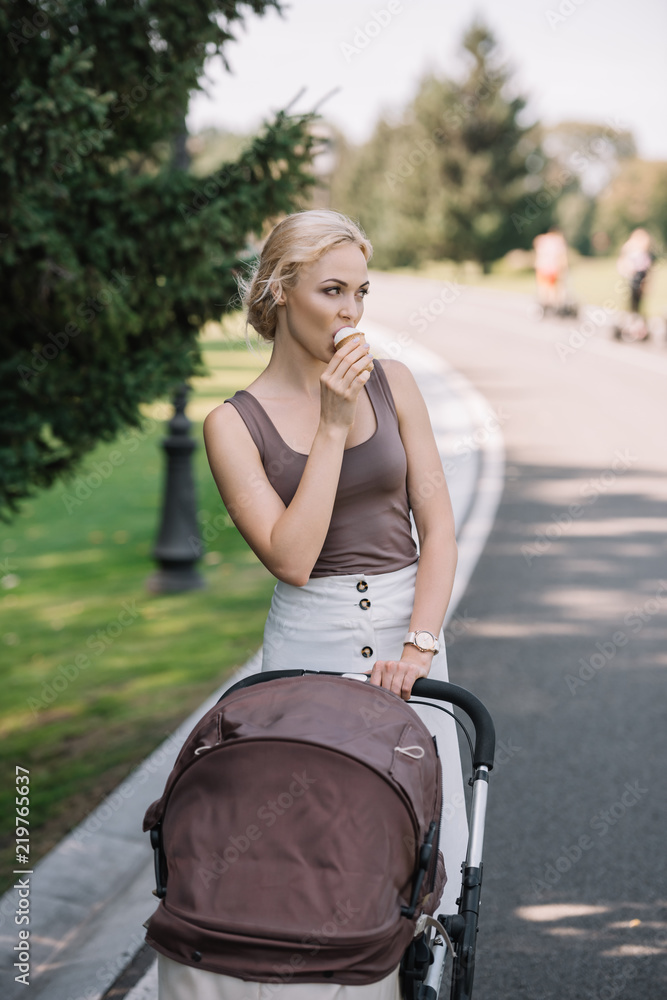 portrait of mother walking with baby carriage and eating ice cream in park portrait of mother walking with baby carriage and eating ice cream in park