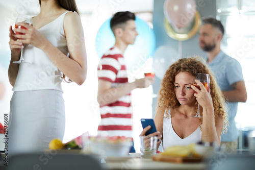 Upset girl having drink by table while reading message or notification in smartphone at party