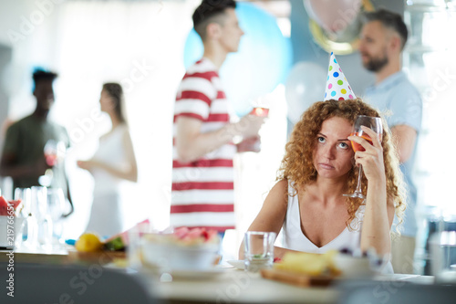Bored girl with glass of drink sitting by table at birthday party while her friends communicating near by