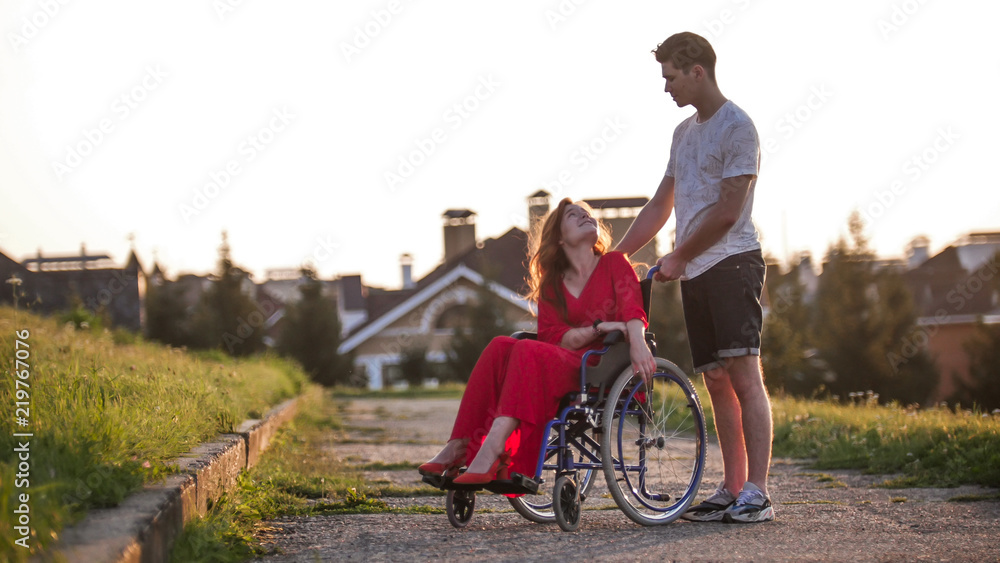 A beautiful girl with red hair in a red dress is sitting in a wheelchair and smiling at her boyfriend who is standing next to her, behind her A beautiful girl with red hair in a red dress is sitting in a wheelchair and smiling at her boyfriend who is standing next to her, behind her