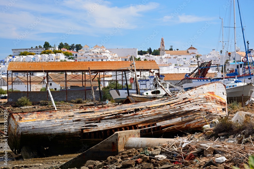Old wooden fishing boat ruin on the riverbank with views towards the town buildings, Ayamonte, Spain.