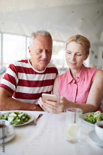 Mature woman with smartphone and her husband communicating through video-chat while sitting by lunch