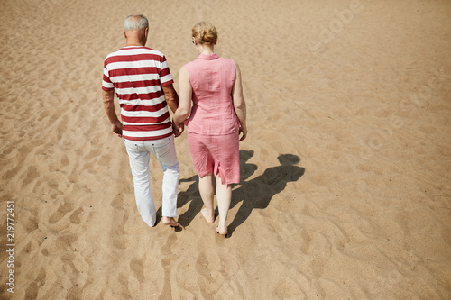 Affectionate senior couple in casualwear walking down sandy beach on hot summer day