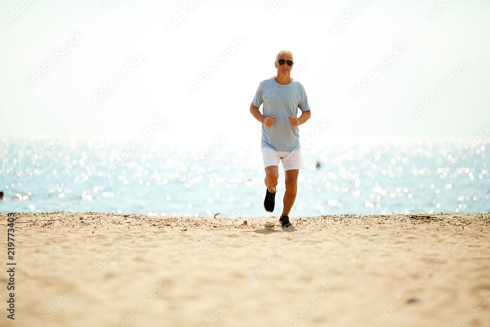 Active pensioner in sportswear running down sandy beach with water on background