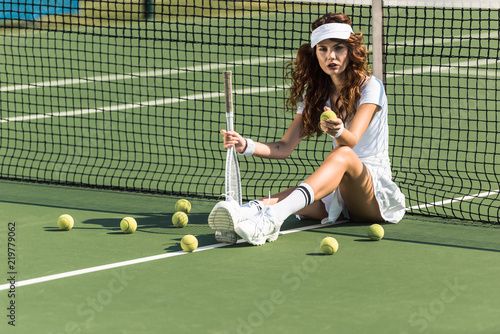 beautiful female tennis player with racket sitting near tennis net on court with tennis balls around © LIGHTFIELD STUDIOS